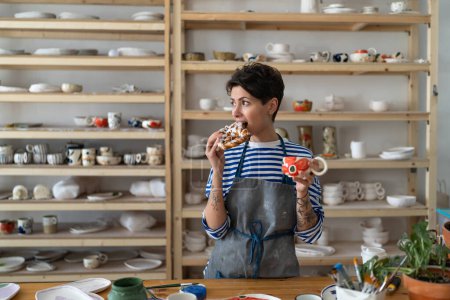 Foto de Relaxed woman in apron drink coffee from handmade ceramic cup and has snack in pottery studio. Tattooed female artist has break at workplace after work. Small business and art concept - Imagen libre de derechos