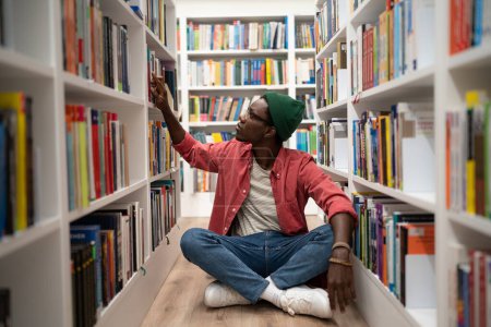 Foto de Autoeducación. Un joven afroamericano se sienta en el piso de la biblioteca entre estanterías, pasando tiempo en la librería, buscando un libro, un estudiante negro preparándose para el examen universitario. Día Mundial del Libro - Imagen libre de derechos