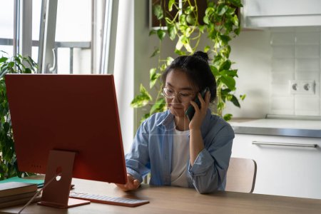 Photo for Enterprising pensive Asian woman sits at computer desk in home office making phone call. Professional casual Chinese girl in search of work calls companies wanting to get job as secretary or clerk - Royalty Free Image
