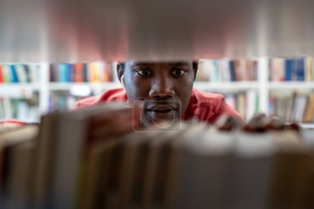 Photo for African American man standing between bookshelves in university library, picking up book from shelf, guy student choosing textbook, looking for literature while preparing for exam. Education - Royalty Free Image