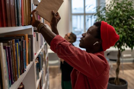 Téléchargez les photos : Concentrated young African American guy picking book for preparing for exam, project in literature or test from bookshelves in university campus. Man college student in library. Education and studying - en image libre de droit