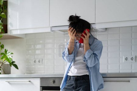 Foto de Worried, anxious young Asian woman holding smartphone receiving bad news from doctor via phone while standing in kitchen at home. Stressed millennial girl talking on cellphone, feeling upset - Imagen libre de derechos