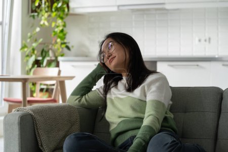Photo for Positive cheerful Asian woman in glasses relaxes sits on sofa enjoying silence and loneliness. Happy carefree Korean girl zoomer smiles leaning head on hand dreaming about future vacation or holidays - Royalty Free Image