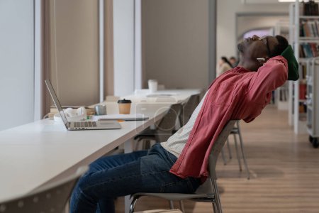 Téléchargez les photos : Jeune étudiant afro-américain épuisé se sentant fatigué de l'apprentissage en ligne, assis au bureau avec un ordinateur portable dans la bibliothèque tenant la main derrière et se reposant des études, prenant une pause pendant les cours en ligne - en image libre de droit