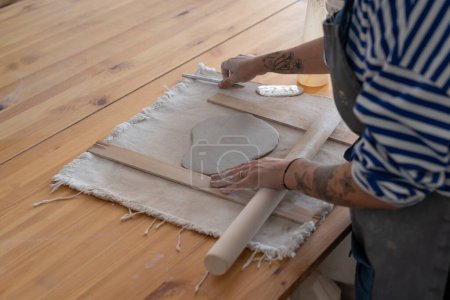 Téléchargez les photos : Woman potter artist in apron use wooden rolling pin to roll out clay making handmade utensils. Process of creating dishes by professional ceramics specialist in workshop with table made of planks - en image libre de droit