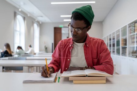 Foto de Talented young black guy scientist wearing glasses studying in library, reading book and taking notes. Focused African American male student writing research summary, sitting at desk. Education - Imagen libre de derechos