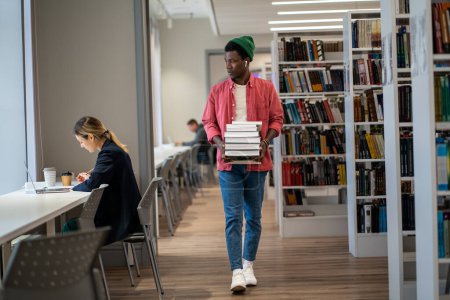 Photo for Bookworm. Full length of African American international student guy walking with stacked books through college library. Male librarian organizing and managing resources. Education concept - Royalty Free Image