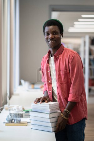 Photo for Young cheerful African American man librarian smiling at camera while standing with pile of books at public library, creating or updating online database, cataloging library materials - Royalty Free Image