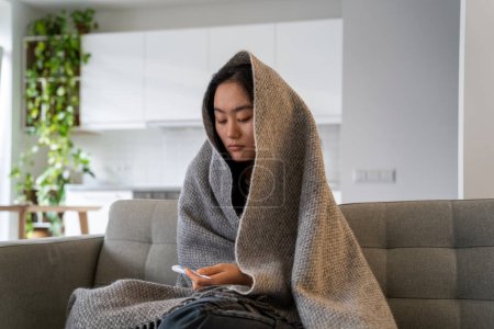 Téléchargez les photos : Mauvaise santé asiatique chinoise jeune femme assise sous une couverture de laine sur un canapé souffrant de froid, regardant thermomètre mal à l'aise avec la fièvre élevée maladie du virus de la grippe migraine de grippe froide. - en image libre de droit