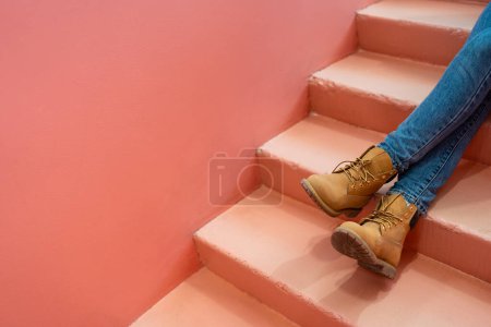 Téléchargez les photos : Legs in blue jeans and light brown boots stretched out on coral-pink staircase against pink-orange wall. Copy space for advertising, to insert text or slogan. Relax comfort design color concept. - en image libre de droit
