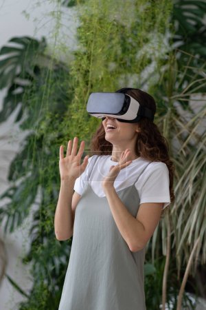 Téléchargez les photos : Jeune femme ravie de passer du temps libre dans la réalité virtuelle tout en se relaxant dans le jardin à la maison, jardinière stupéfaite jouant à des jeux VR, en utilisant la technologie interactive. Concept de cyberespace. - en image libre de droit