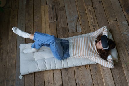 Téléchargez les photos : Femme caucasienne détendue dans le casque de lunettes de réalité virtuelle couché sur le repos au sol en regardant la vidéo 3d ar utiliser la technologie moderne augmentée futuriste vr lunettes. Jeune dispositif de test féminin occasionnel pour la vidéo - en image libre de droit