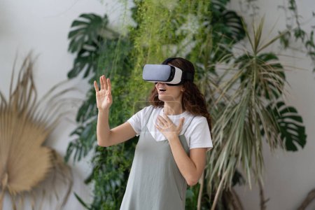 Téléchargez les photos : Animation virtuelle à la maison jardin. Femme heureuse émotionnelle dans le casque vr en utilisant VR casque, jouer à des jeux vidéo interactifs, explorer la réalité augmentée sont excités choqués par l'interface. Cyberespace. - en image libre de droit