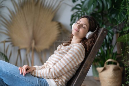 Photo for Young woman sitting in armchair in cozy home garden, dreaming, resting, listen to music on wireless headphones. Female wear earphones, closed eyes and relaxing surrounded by houseplants in living room - Royalty Free Image