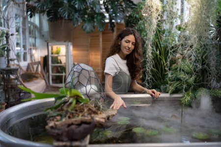Photo for Young gardener woman relaxing after work, touches with hand water in freestanding bathtub with an aquatic houseplant in cozy green house. Greenery at home garden, love for plants, hobby concept - Royalty Free Image