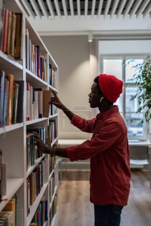 Téléchargez les photos : Black student man in red hat stands between library librcases, African American guy pick up book from shelf while studying in university library, using learning materials. Possibilités d'éducation - en image libre de droit