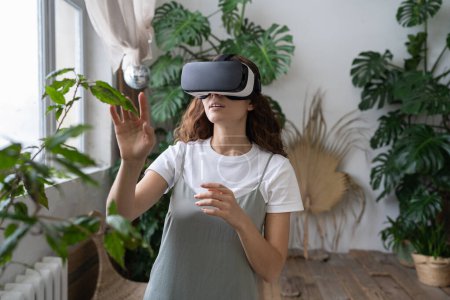 Foto de Jardinería casera, tecnología virtual, concepto metaverso. Mujer joven hispana tocando planta de interior en el espacio inmersivo VR. Mujer italiana usando gafas VR en invernadero. Realidad aumentada. - Imagen libre de derechos