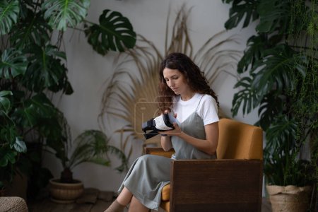 Téléchargez les photos : Curieux jeune femme tenant casque VR assis dans un jardin familial confortable avec plante tropicale d'intérieur Monstera deliciosa, designer paysagiste test moderne achat innovant lunettes de réalité virtuelle - en image libre de droit