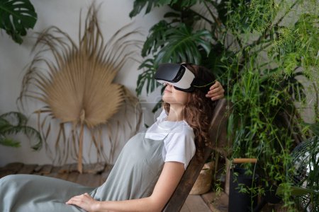 Téléchargez les photos : Femme hispanique incroyable en réalité virtuelle lunettes reposant dans un environnement virtuel relaxant au jardin à la maison, assis dans un fauteuil avec les mains derrière la tête. AR, concept de cyberespace. Fille portant un casque VR. - en image libre de droit