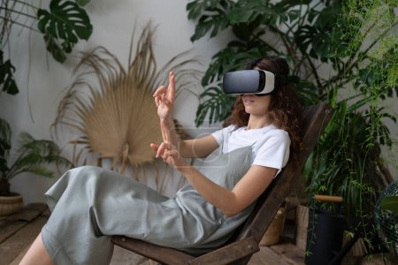Téléchargez les photos : Millennial girl in vr lunettes tactile écran interactif dans l'air utiliser la technologie de réalité virtuelle pour le divertissement à la maison dans le jardin de la maison confortable. Les jeunes femmes portent des lunettes de casque portable jouer au jeu vidéo - en image libre de droit