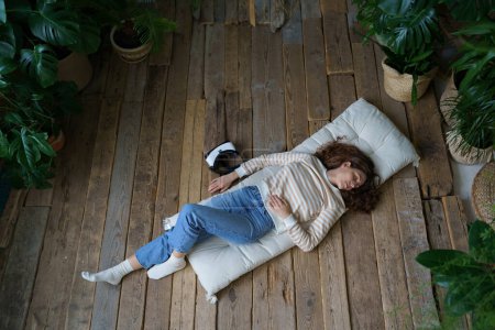 Photo for Relaxed italian woman with virtual reality glasses headset lying on wooden floor at home, rest after watching 3d ar video. Calm young casual female sleeping on mattress surrounded by tropical plants. - Royalty Free Image