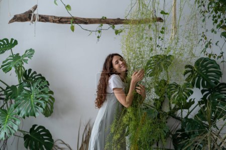 Téléchargez les photos : Young Spanish girl stands among green plants on stepladder and hugs asparagus in living room, monstera on background. Home comfort harmony. Hobby, houseplant lovers, gardening concept - en image libre de droit