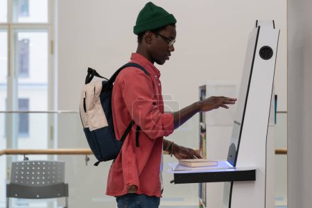 Photo for Young African student guy with backpack standing in library using self-service terminal to borrow or return books, searching literature browsing catalogue, touching screen of self-checkout kiosk - Royalty Free Image