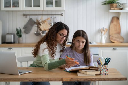 Photo for Nervous emotional mother helps daughter prepare report for school sits with textbook at kitchen table. Confused girl teenager reads books and makes notes in workbook under supervision of woman tutor - Royalty Free Image