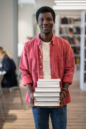 Photo for Portrait of happy African American student guy holding pile of books smiling at camera while standing at library, enjoying studying and University life, selective focus. Education and self-study - Royalty Free Image