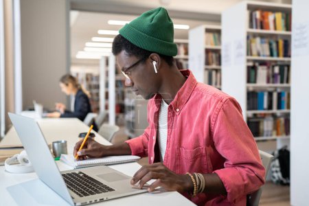 Foto de Estudiante afroamericano enfocado que aprende en línea tomando notas durante el webinar, estudiando remotamente en la biblioteca. Joven chico negro viendo vídeo educativo o conferencia en el ordenador portátil. Concepto de estudio remoto - Imagen libre de derechos
