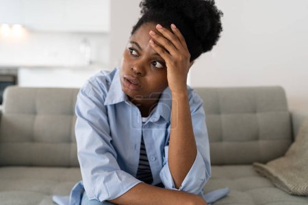 Photo for Flustered African American woman very nervous sighing and touching face with hands sits on couch of home. Young black girl nervous, thinking, feeling fear after wrong decision or problems at work. - Royalty Free Image