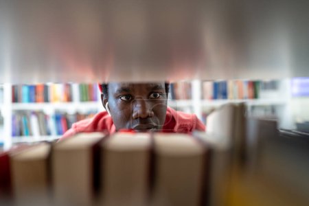 Téléchargez les photos : Gros plan sur le visage concentré d'un étudiant afro-américain entre deux bibliothèques universitaires. Focalisé noir jeune homme choisir littérature vérifier l'information en librairie pour des études universitaires efficaces - en image libre de droit
