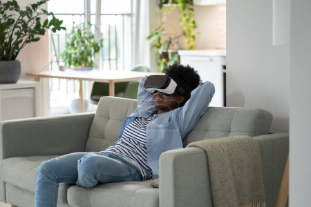 Téléchargez les photos : Impressions smiling African woman using virtual reality headset immersing herself in metaverse. Happy satisfied black girl sitting on couch with hands behind head experiencing VR and AR technology - en image libre de droit