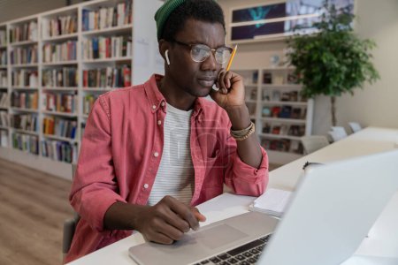 Photo for Focused on screen black guy student search new idea inspiration in campus library. Concentrated African American freelancer man hold pencil contemplates solution of task sits at work table with laptop - Royalty Free Image