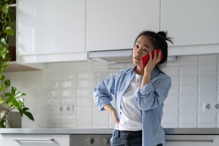 Foto de Joven mujer asiática decepcionada de pie en la cocina en casa hablando en el teléfono inteligente escuchar malas noticias. Mujer estresada sosteniendo el teléfono móvil sintiéndose molesta durante una conversación telefónica desagradable - Imagen libre de derechos