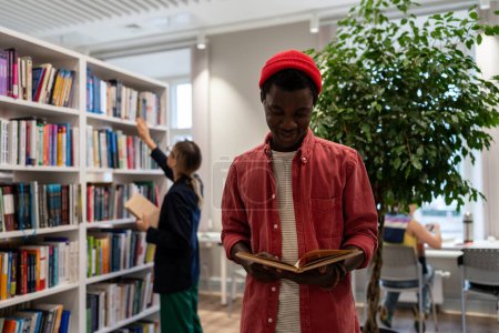 Téléchargez les photos : Joyeux Afro-Américain debout sur le campus universitaire lire acquérir de nouvelles compétences, améliorer les connaissances en langues étrangères sur fond bibliothécaire organise des livres correctement sur étagères formulaires commandes pour les étudiants - en image libre de droit