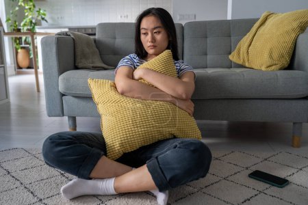 Photo for Upset korean girl feels lonely sadness suffering from bad relationship. Unhappy asian woman with pillow in hands thinks mental troubles. Depressed young female crushed by breakup sits on floor at home - Royalty Free Image