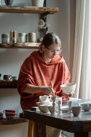 Photo for Creative handcraft clay study master class for video blog. Puzzled focused professional craft young woman painting on ceramic bowl. Modern potter workshop invent with new idea for future earthenware. - Royalty Free Image