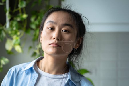 Photo for Close up portrait of young pensive serious Asian woman looking at camera while standing in kitchen at home, thinking about problems or unpleasant things, feeling down. Korean beauty standards - Royalty Free Image