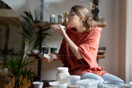 Photo for Sculptor artisan girl blows dust off clay item in cozy comfortable workshop studio. Careful production of designer earthenware before glassing or kilning. Potter making ceramic saucer for custom order - Royalty Free Image