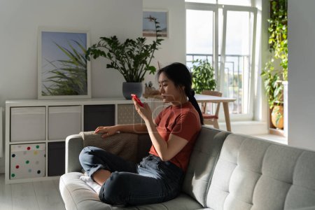 Téléchargez les photos : Jeune Chinoise asiatique détendue assise sur un canapé à la maison, bavardant sur un téléphone portable, faisant du shopping sur le site de surf défilant en ligne, choisissez une vidéo à regarder. Procrastination due à la dépendance aux médias sociaux - en image libre de droit