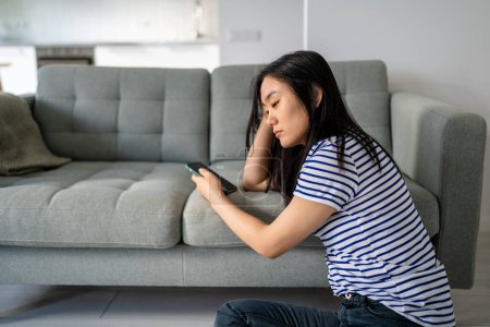 Downcast mujer asiática mirando tristemente a la pantalla del dispositivo de lectura de mensaje desagradable o a la espera de la llamada. Molesto chica china usando el teléfono celular a regañadientes leer noticias ponderando problema considerando soluciones.