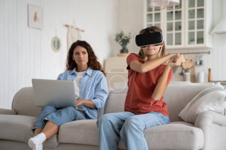 Téléchargez les photos : Adolescente enfant portant des lunettes de réalité virtuelle jouant à des jeux de réalité virtuelle pendant que la mère travaille à distance, assise ensemble sur le canapé à la maison, mère parent et enfant passent du temps avec des gadgets - en image libre de droit
