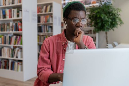 Photo for Focused pensive African American hipster freelancer man in eyeglasses working at laptop at library workplace table thinking over IT online startup project. Concentrated black guy student solves task. - Royalty Free Image