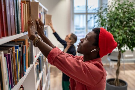 Photo for Puzzled serious on finding right educational literature for difficult project student looks through bookshelves in library. African American guy search for book title picks one for lecture class task. - Royalty Free Image