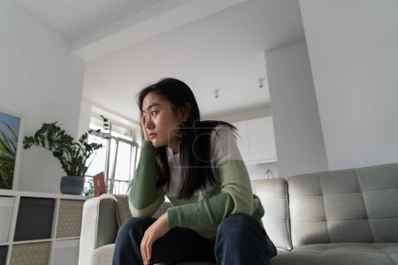 Photo for Unmotivated pensive Asian student girl sits on couch leaning head on hand. Procrastinating Chinese woman is stressed out because she doesnt want to start preparing for university entrance exams - Royalty Free Image
