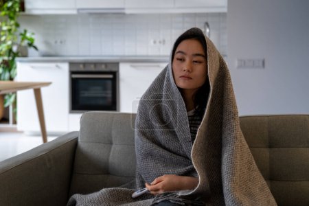 Photo for Sick exhausted Asian woman with thermometer to measure body temperature sit on sofa at home wrapped in blanket. Suffering Chinese girl experiences health problems due to abnormally cold winter weather - Royalty Free Image