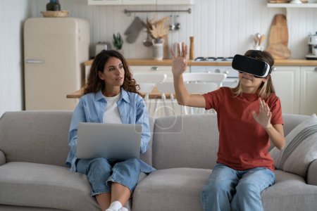 Photo for Interested teenage girl uses VR headset to visit metaverse or watch 3D virtual reality movies from comfort home. Progressive European schoolgirl waving hands sits on sofa near mother woman with laptop - Royalty Free Image