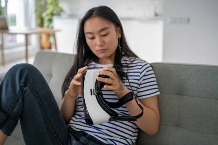 Photo for Focused Chinese woman sits on couch considers 3D virtual reality glasses for immersion in gaming universe. Concentrated Asian young female resting at home choosing new activity ready to use VR headset - Royalty Free Image