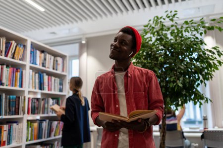 Photo for Satisfied African American student stands in library with open book looks dream about passed exam enjoy interesting effective studies in higher school. Successful excellent guy learner get scholarship - Royalty Free Image
